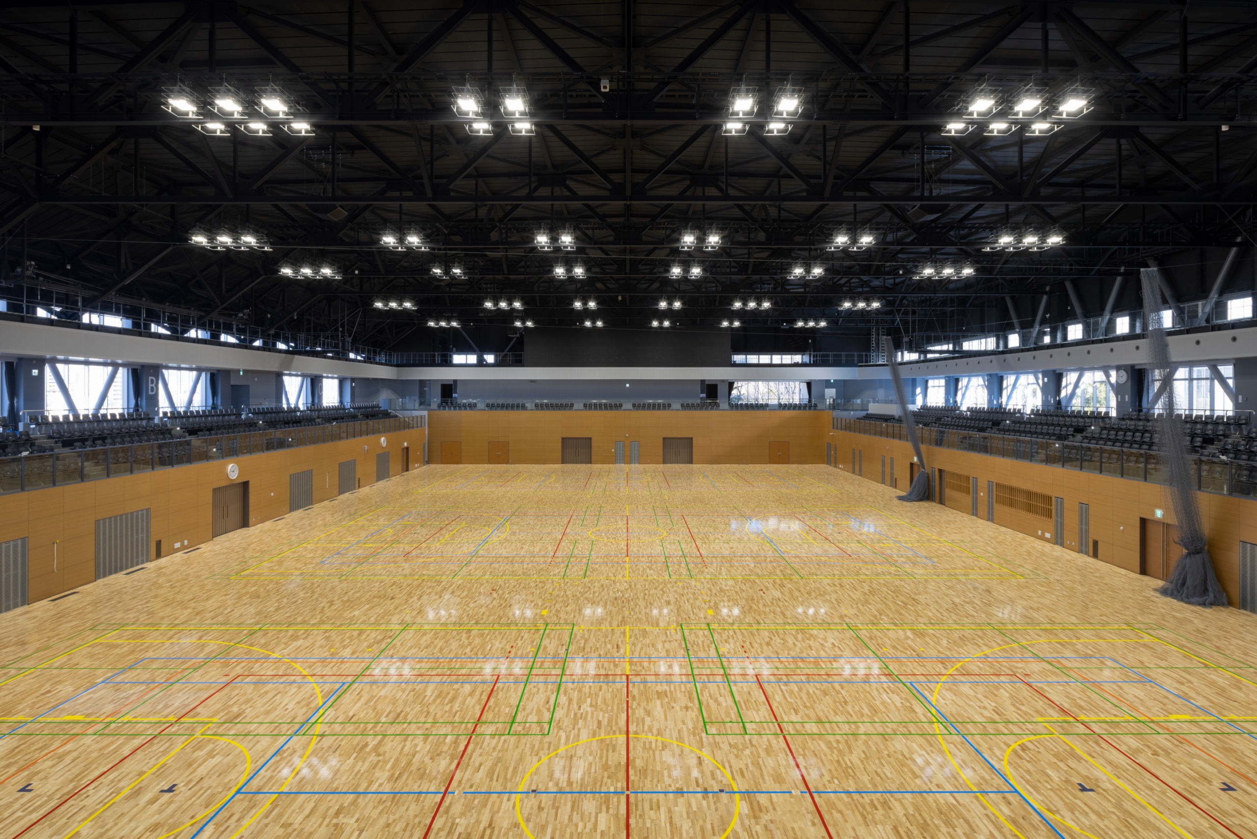 沼津市総合体育館メインアリーナの広々とした全景。明るいLED照明が多数設置された高い天井と、複数の競技ラインが引かれた美しい木目調のフロアが特徴。大規模なスポーツ大会や合宿に最適な観客席も見える屋内施設。