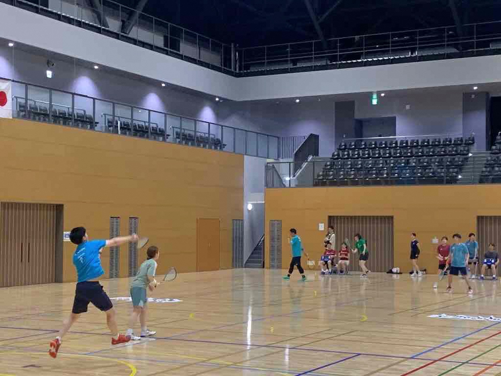 沼津市総合体育館の多目的アリーナでクロスミントンをプレーする選手たち。高速で飛び交うスピーダーを追いかけ、集中してフットワークを使う様子が捉えられている。