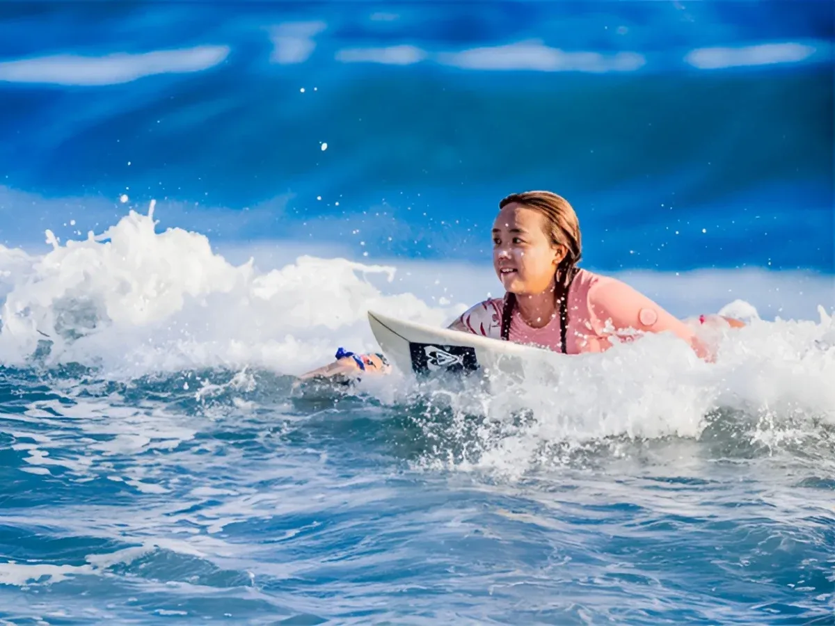 ピンク色のウェアを着用し、波打ち際の白い泡に囲まれながらサーフボードの上で集中した表情を見せる佐藤李選手。