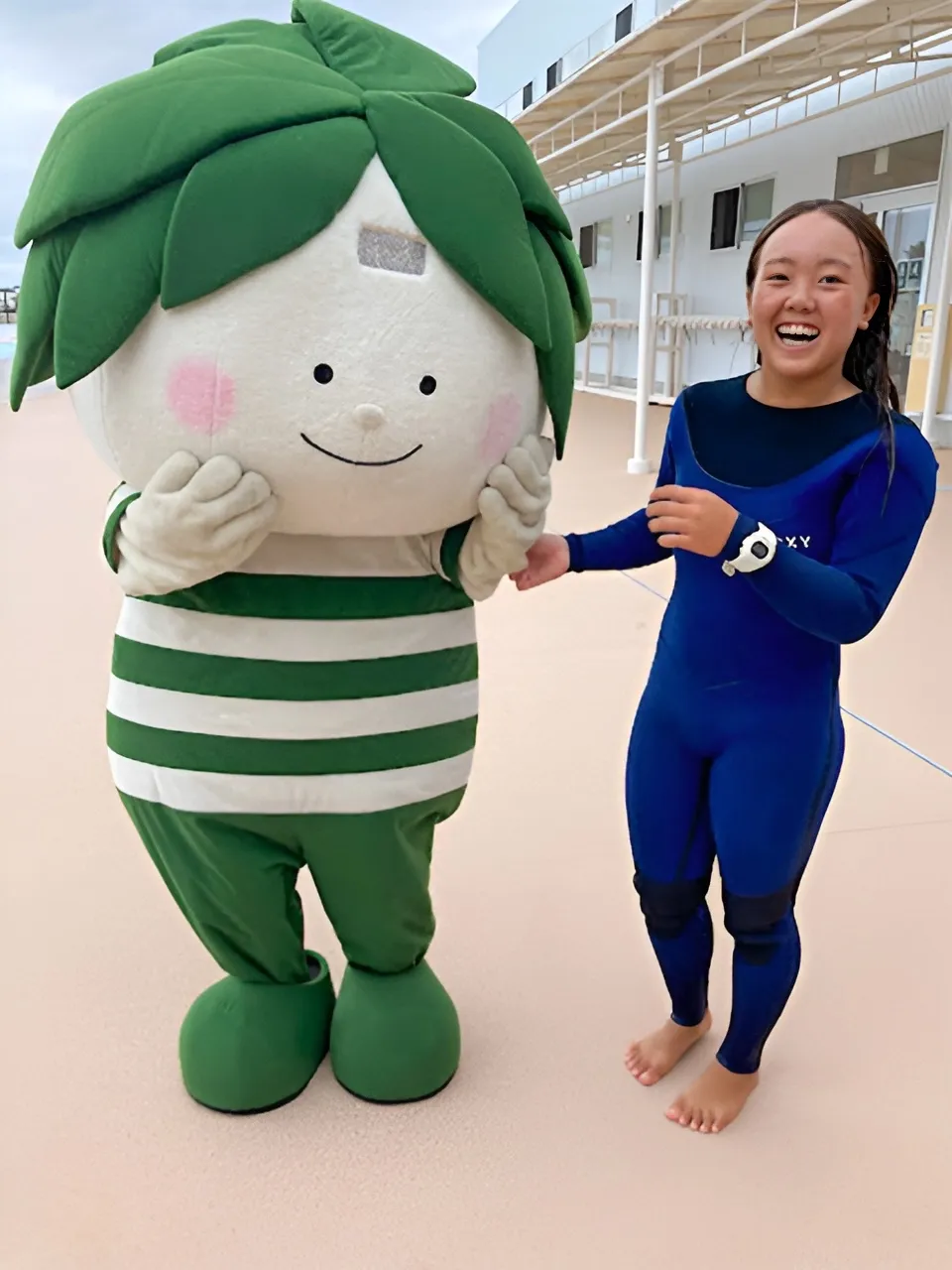 青いウェットスーツ姿の佐藤李選手が、マスコットと笑顔で触れ合っている様子。