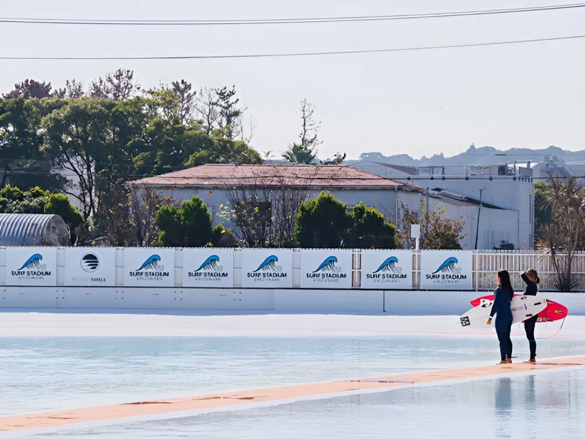静波サーフスタジアムのプールサイドに立ち、サーフボードを抱えて水面を見つめる佐藤李選手の背後からのカット。